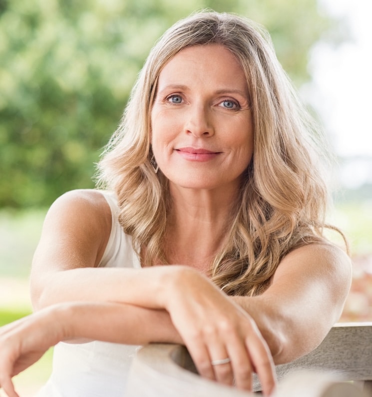 Happy senior woman relaxing on bench in the lawn. Close up face of a mature blonde woman smiling and looking at camera. Retired woman in casuals sitting outdoor in a summer day. (Happy senior woman relaxing on bench in the lawn. Close up face of a mat